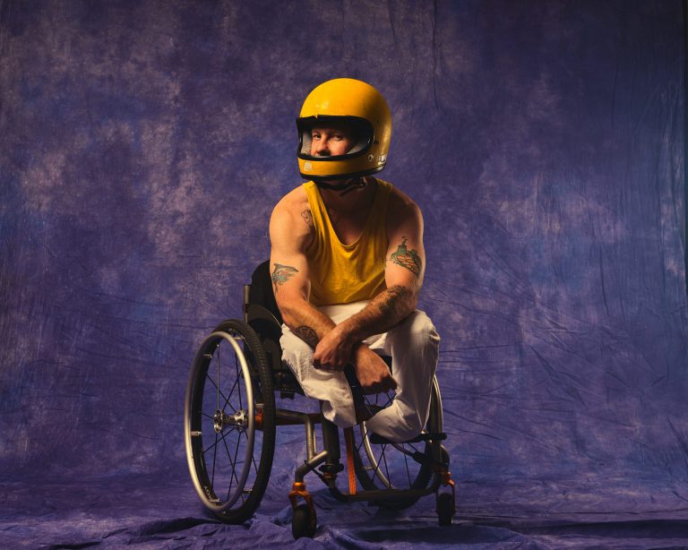 A man with a disability sits in a wheelchair, flexing his arms, dressed in a yellow helmet and singlet and beige pants