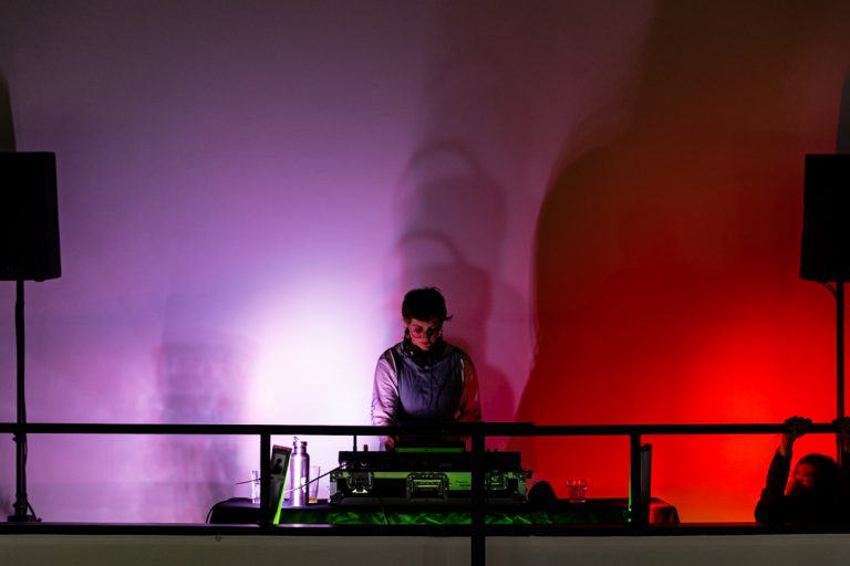A person standing in front of their DJ desks against a backdrop of purple and red light